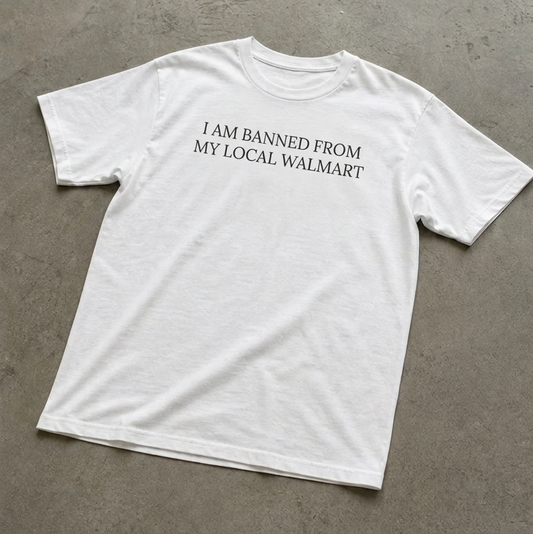 White t-shirt with text 'I AM BANNED FROM MY LOCAL WALMART' on a concrete floor.